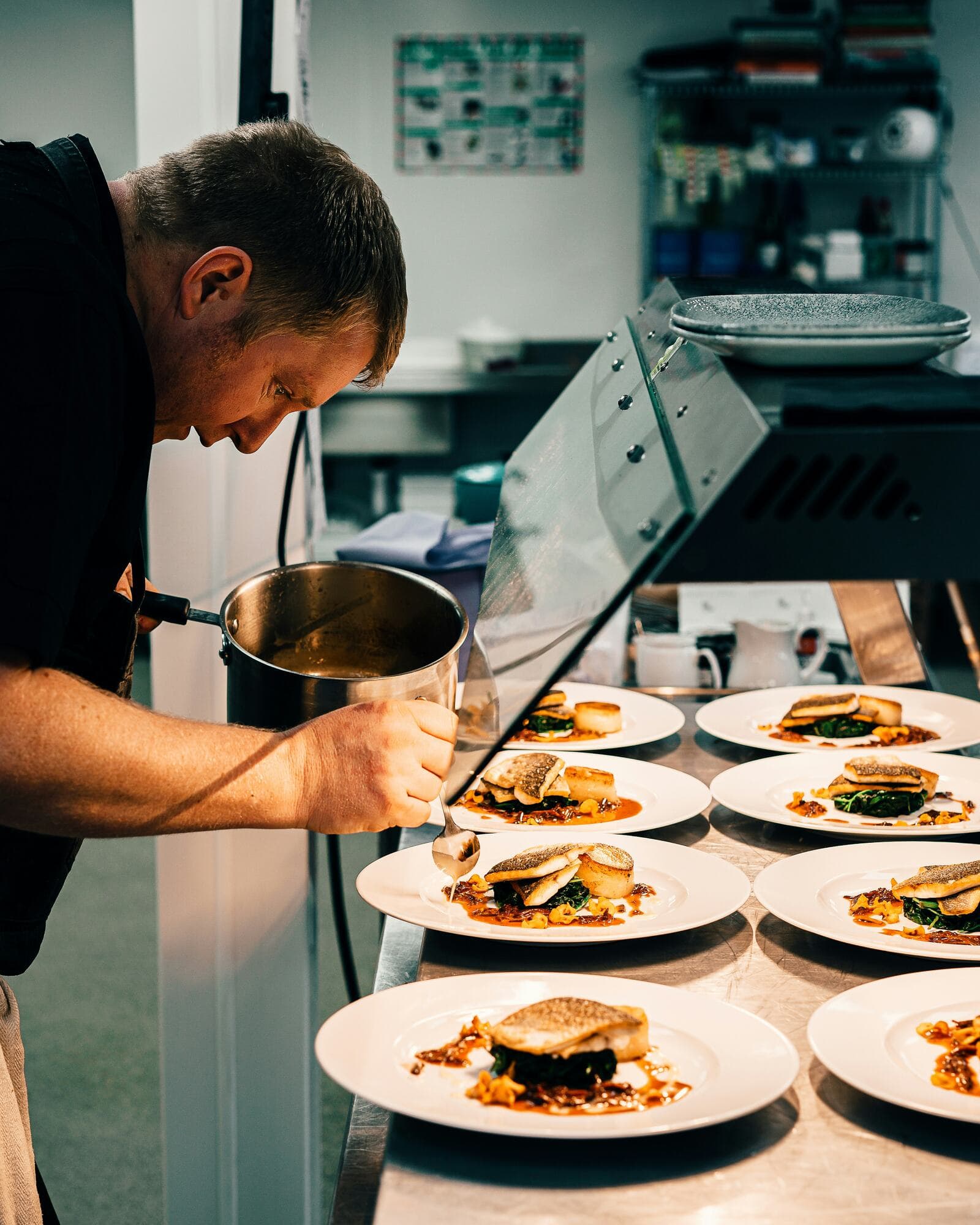 A cook leaning over a row of plated fish dishes, pouring sauce from a steel pot