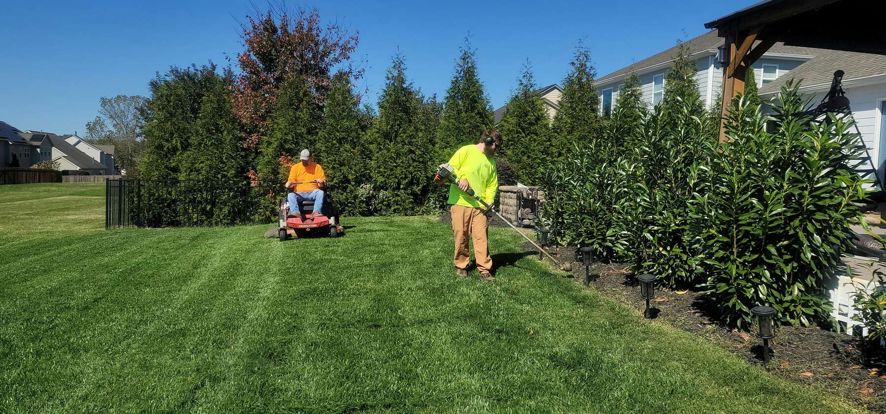 Two landscapers on a suburban lawn, one riding a zero-turn mower and one edging the beds with a string trimmer