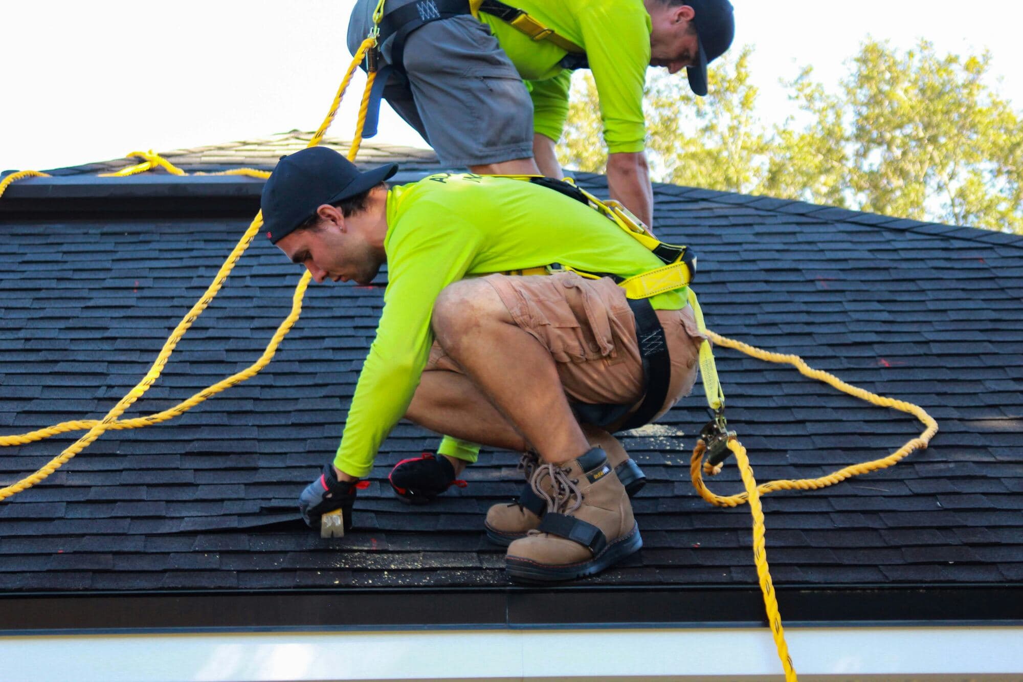 Two roofers in safety harnesses and bright green shirts working on an asphalt shingle roof, one crouched over the shingles with a hand tool